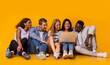 © Prostock-studio - Carefree international teenagers sitting on floor and using laptop, yellow studio background, panorama