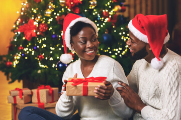 Wall Mural - Loving black guy surprising his girlfriend with Christmas gift at home, celebrating winter holidays together