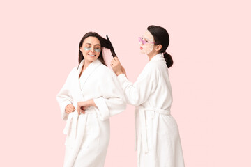 Poster - Young women in bathrobes with facial mask and under-eye patches doing hair on pink background