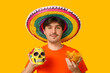© Pixel-Shot - Portrait of young man with sombrero, tortilla chips and painted human skull on yellow background. National Tortilla Chip Day celebration