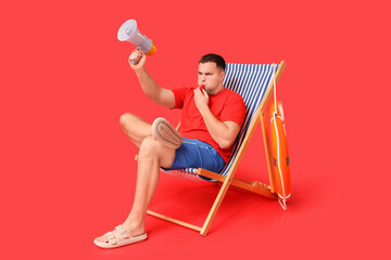 Wall Mural - Young lifeguard with lifebuoy and megaphone sitting on beach lounge and whistling against red background