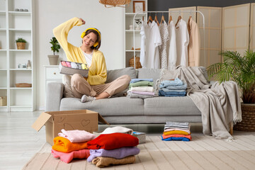 Wall Mural - Young woman in headphones with stacks of clothes on sofa near donation cardboard box at home