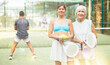 © JackF - Young and elderly female tennis players standing on outdoor court after game on summer day, holding balls and rackets for padel in hands..
