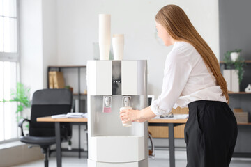 Wall Mural - Businesswoman pouring water from cooler in office, back view