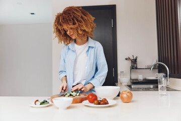 Wall Mural - Woman preparing healthy meal in modern kitchen with curly hair, cutting vegetables on wooden board