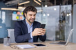 © Liubomir - Businessman in a suit smiling while using a smartphone in a modern office, surrounded by a laptop and tablet, showcasing digital communication and multitasking.
