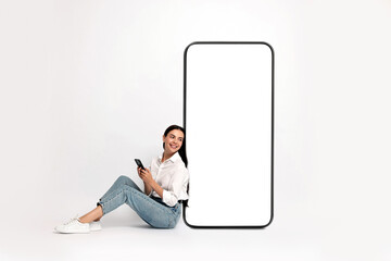 Wall Mural - Beautiful young woman with smartphone sitting near big mobile phone with blank screen on light background