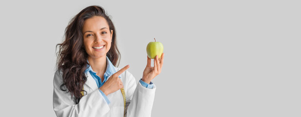 A health professional woman wearing a white lab coat is smiling widely while pointing at a fresh green apple. The setting appears to be a laboratory or health-focused environment, copy space