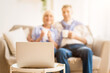© Prostock-studio - Senior couple watching tv online on laptop and drinking tea on comfortable sofa, focus on computer