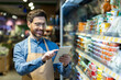 © Liubomir - Confident grocery store worker uses digital tablet while standing in supermarket aisle. Surrounded by various products, he engages with technology to manage inventory.