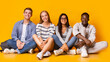 © Prostock-studio - Happy multiracial students sitting on floor and posing over yellow studio background, students lifestyle concept