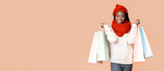 A joyful young African American woman stands smiling while holding several colorful shopping bags. She is dressed warmly in a cozy sweater and a bright red winter hat, copy space