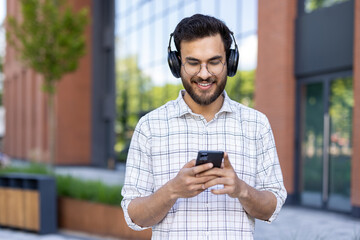 Wall Mural - A smiling young Indian man in headphones is walking on a city street and using a mobile phone