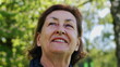 © Marco - Elderly woman looking up with a smile in a park, surrounded by vibrant greenery. The sunlight illuminates her face, highlighting her joyful expression