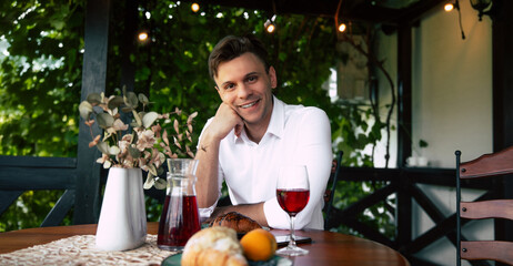 Close up portrait of young handsome man smiles while seated at a table with a pastry, a glass of drink, and fresh flowers in a bright café outdoors.