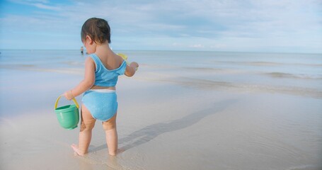 A young toddler child in a blue swimsuit playing with sand toys bucket and shovel on a sandy tropical beach the ocean and sky in the background summertime