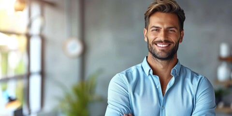 Smiling young man in blue shirt stands confidently with arms crossed, exuding positivity and professionalism in modern office environment, showcasing approachable business demeanor.