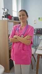 Wall Mural - A young woman in pink scrubs with a stethoscope stands confidently with crossed arms in a clinic room.