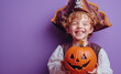 © netrun78 - A joyful boy dressed in a pirate costume laughs gleefully while holding a carved Halloween pumpkin, set against a vibrant violet backdrop.