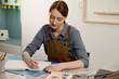 © pressmaster - Young creative craftswoman in apron sitting by desk with art supplies and sketching with pencil on page of open notepad in studio