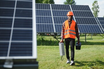 Wall Mural - Portrait of young handsome African American craftsman in protective helmet. Man in uniform and with tools standing among solar panels