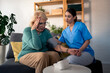 © Dorde - A female nurse in scrubs assists an elderly woman with her blood pressure check, providing medical care in a home setting, with a concerned look from the patient.