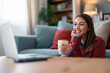 © Dorde - A smiling woman enjoys a video conversation, comfortably settled in her home's living room. Her casual attire and mug suggest a relaxed vibe during remote communication.