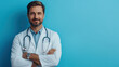 © khonkangrua - Portrait of a confident male doctor with a stethoscope around his neck, standing against a blue background, arms crossed.