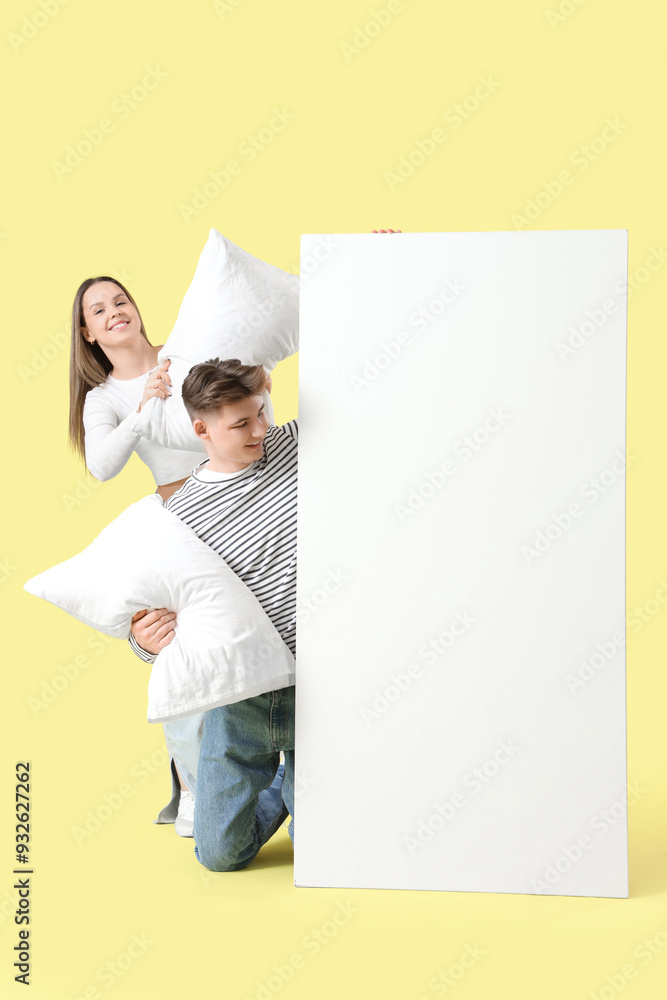 Young couple with pillows and blank poster on yellow background
