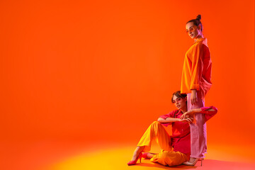 Wall Mural - Beautiful young fashionable sisters in stylish outfits sitting on orange background