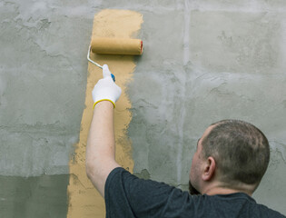 Wall Mural - A man in a black shirt and gloves paints a concrete wall with a roller, using light brown paint