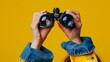 © Farid - female hands hold black binoculars on a bright yellow background.