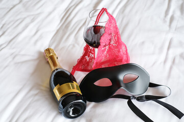 Romantic sex date, red lace panties, black mask and wine glasses on the bed. Concept of night of passion, female lingerie. Woman accessories.