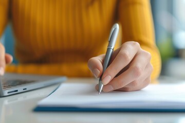 Close-up of a woman writing notes with a pen next to a laptop, symbolizing work, study, or planning activities.