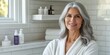 © mashimara - Elegant mature woman with gray hair in a bathrobe, standing by a window in a bathroom with white subway tiles, exuding confidence and grace.