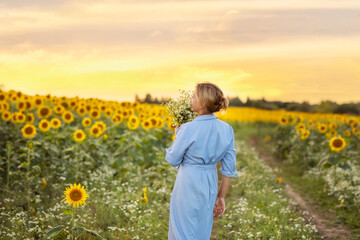 happy woman with a bouquet of daisies walking along a dirt road in a field of sunflowers