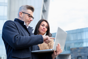 Business partners discussing project outside office, focused on laptop, collaborating for success in urban setting. Dynamic teamwork in digital age