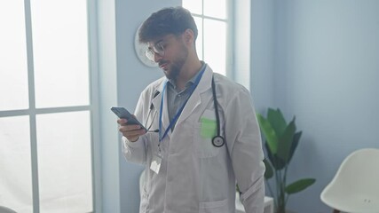 Poster - A young bearded man in a doctor's coat with a stethoscope, using a smartphone in a bright hospital room.