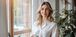 © Jirawatfoto - A woman in white shirt smiles softly while holding cup by window, surrounded by plants.