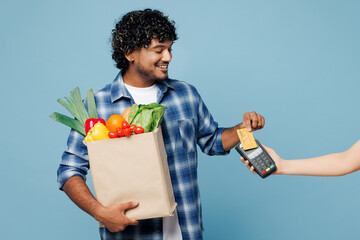 Wall Mural - Young Indian man wear shirt hold craft bag for takeaway mock up with food products bank payment terminal process acquire credit card isolated on plain blue background Delivery from shop or restaurant