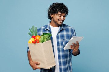 Wall Mural - Young happy Indian man wears shirt hold brown craft bag for takeaway mock up with food products read to do list in notebook isolated on plain blue background. Delivery service from shop or restaurant.