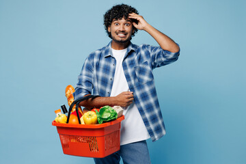 Wall Mural - Young shocked excited surprised Indian man wear shirt hold red basket with food products vegetables put hand on head isolated on plain pastel blue background. Delivery service from shop or restaurant.