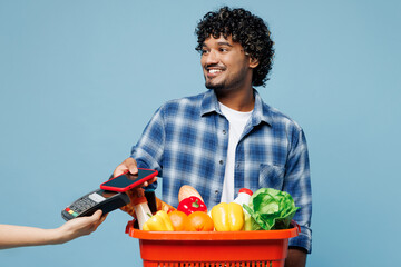 Wall Mural - Young smiling happy Indian man he wear shirt hold red basket with food products, ireless bank payment terminal use mobile cell phone isolated on plain blue background Delivery from shop or restaurant