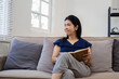 © wichayada - Elderly Woman Relaxing on Sofa at Home, Reading a Book, Enjoying Peaceful Afternoon in Cozy Living Room