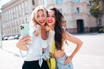 Wall Mural - Two young beautiful smiling female in trendy summer clothes. Sexy carefree women posing in the street. Positive models having fun, hugging in sunny day. With bright makeup, take selfie