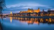 A panoramic shot of Wawel Castle in Krakow during twilight, with the castle glowing against the backdrop of a deepening blue sky and the tranquil Vistula River.