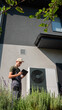 © StockMediaSeller - A technician adjusting a heat pump unit using a tablet in the garden of a modern home, surrounded by lush greenery on a sunny day.