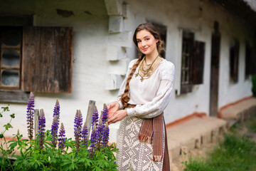 Beautiful blonde long braided hair girl in Ukrainian traditional dress  posing near old houses on nature . Portrait of young smiling attractive woman in accessories  .