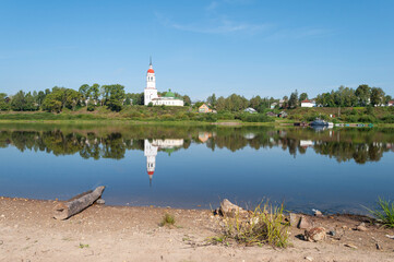 Sukhona river in ancient russian town of Totma, calm water with reflection of ancient church on the other bank. Old log lies in the water, early summer morning