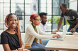 © Katsiaryna - young African American woman in casual clothes posing for the camera while sitting at the table against the background of her colleagues